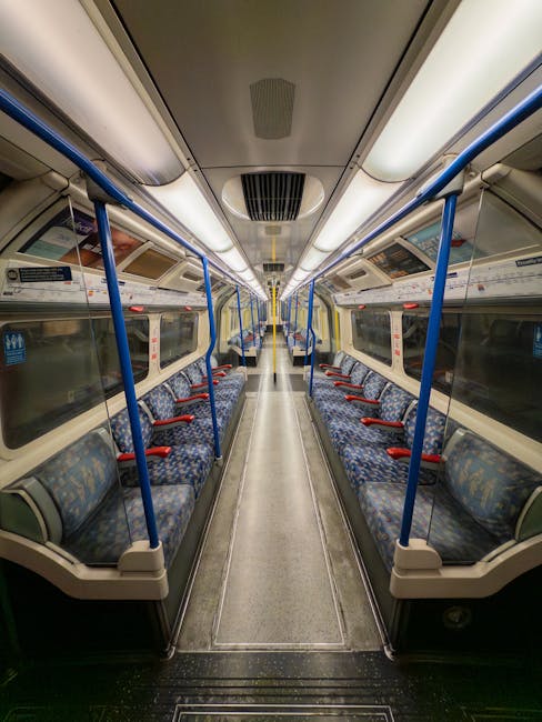 Interior of a public train carriage equipped with patterned fabric-covered seats featuring red armrests and blue metal handrails running along both sides. The floor is made of light-colored, speckled 