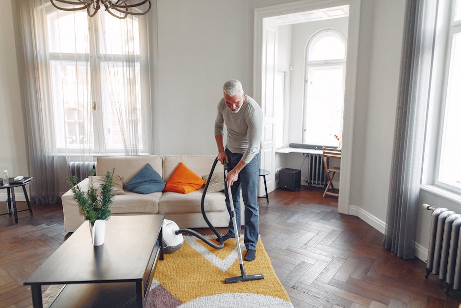Close-up image of a modern vacuum cleaner with a clear plastic dust container filled with debris, connected to a flexible, patterned hose attached to a cleaning floor head resting on a pinkish-beige c